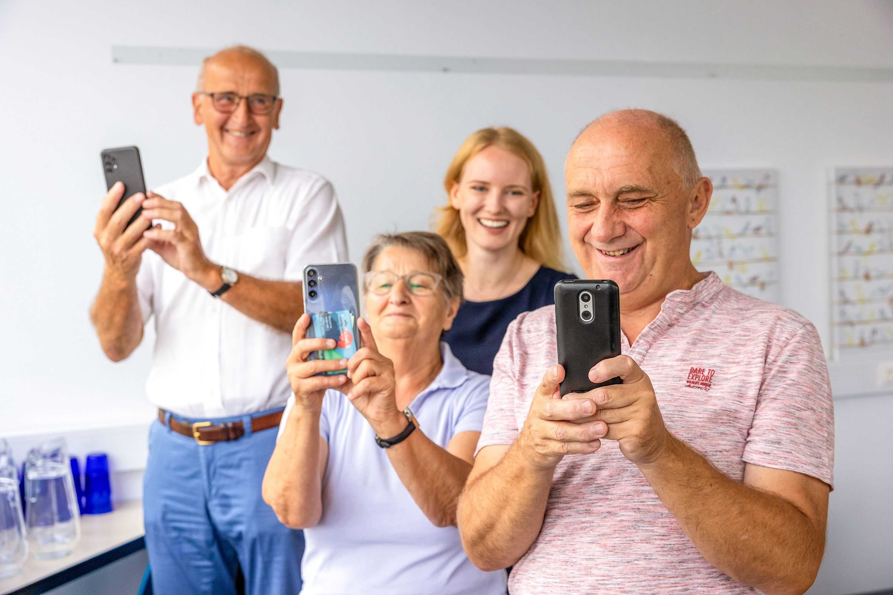 Seniorin und zwei Senioren mit Smartphone in der Hand bei einem Angebot des Salzburger Bildungswerkes, Digitaltrainerin im Hintergrund, alle lachen 